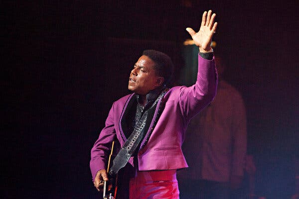 A man in a fuchsia jacket holds a guitar in one hand and raises his other.